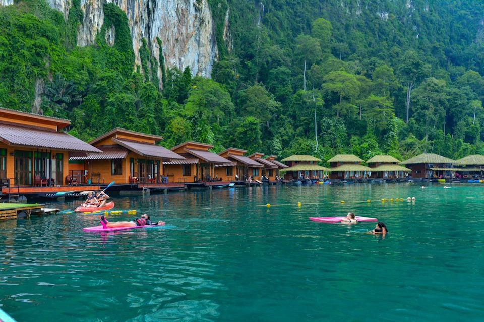 People enjoy relaxing on paddle boards in a turquoise lake near floating wooden cabins, surrounded by lush green cliffs.