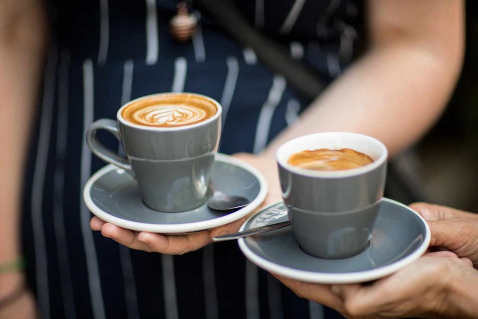 A person holds two gray ceramic cups, each on a saucer, containing latte art and an espresso.