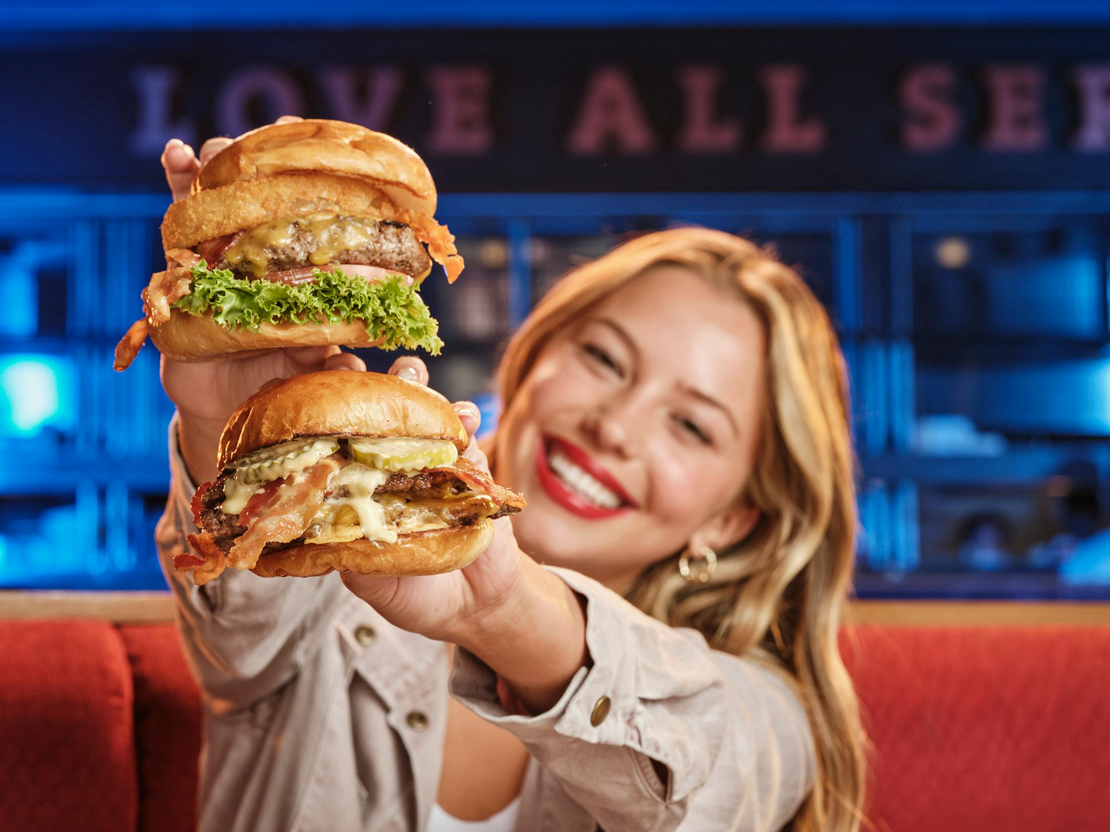 Eine lächelnde Frau, die zwei große Burger in beiden Händen hält, mit einem Restaurantinterieur im Hintergrund.