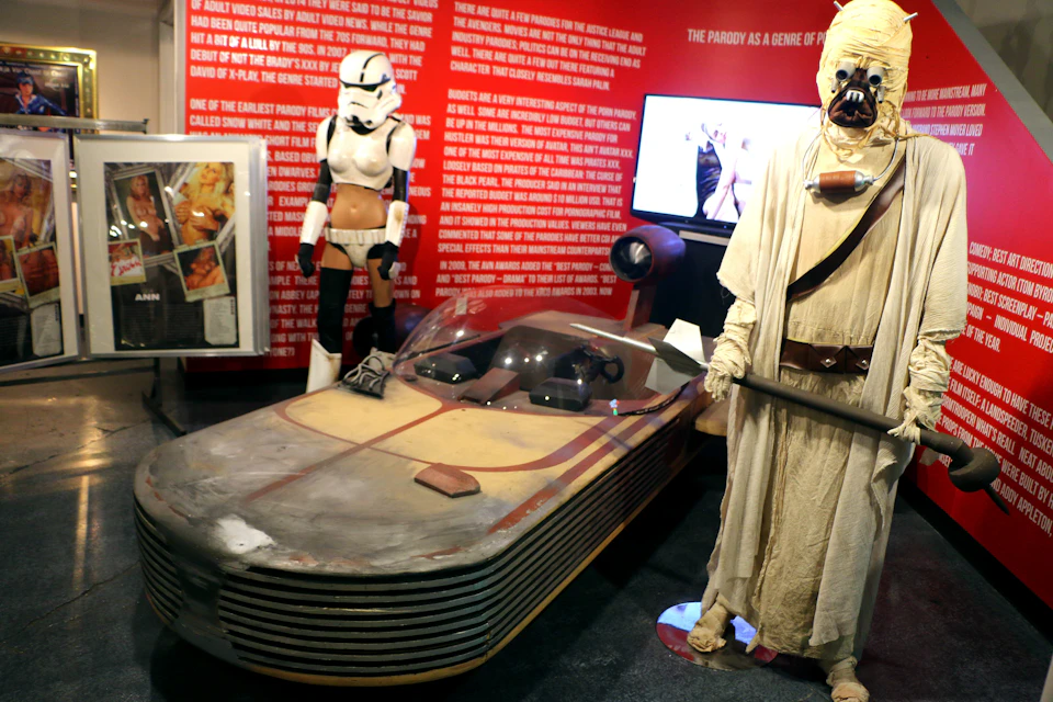 Museum display with a Star Wars landspeeder model, Stormtrooper costume, and Tusken Raider figure. Red wall with text background.