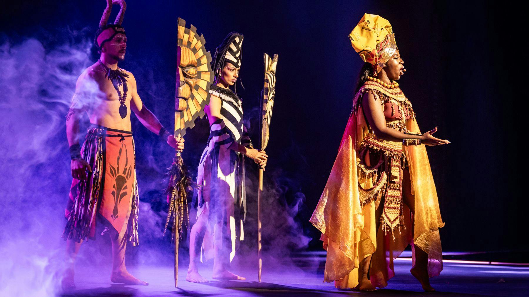 Um artista em palco com um elaborado fato amarelo e laranja com um toucado, acompanhado por outro com um fato com padrão de zebra.