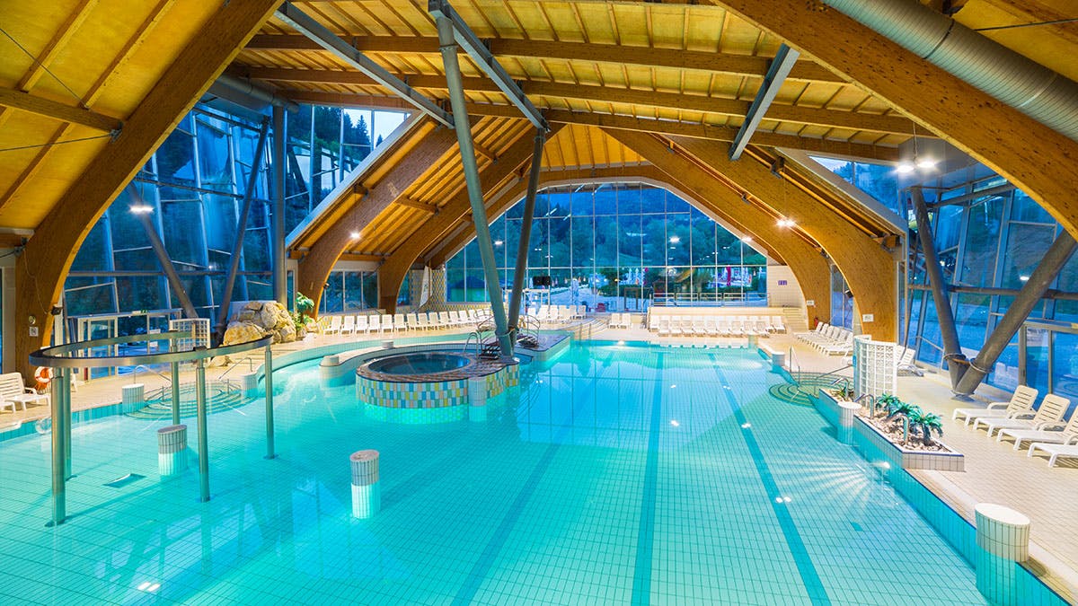 Krytý bazén s dřevěnou architekturou, vířivkou a lehátky, osvětlený teplým osvětlením.