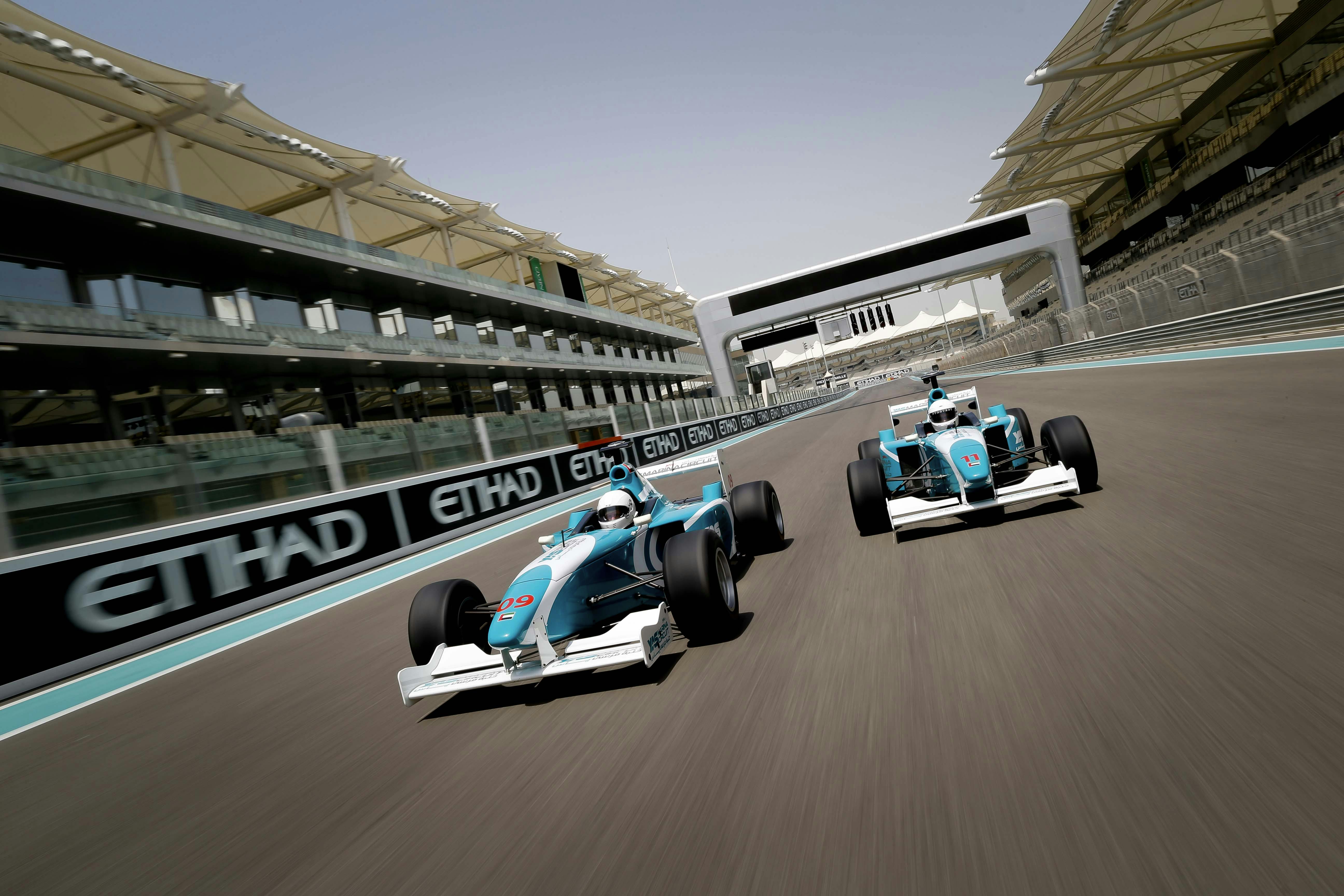 Two blue and white race cars drive side by side on a modern racetrack lined with Ethiad banners and overhead structures.