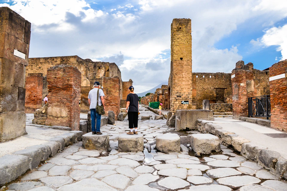 Two people stand on a stone path among ancient ruins with brick and stone structures under a partly cloudy sky.