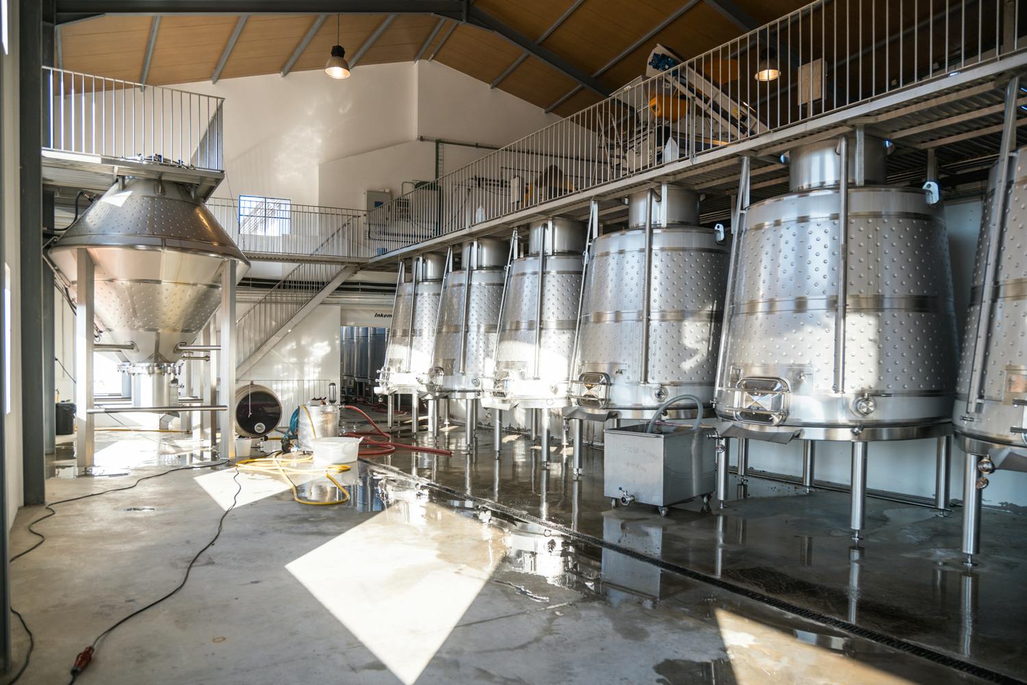Een moderne wijnmakerij met roestvrijstalen fermentatietanks, een tussenverdieping en heldere verlichting.