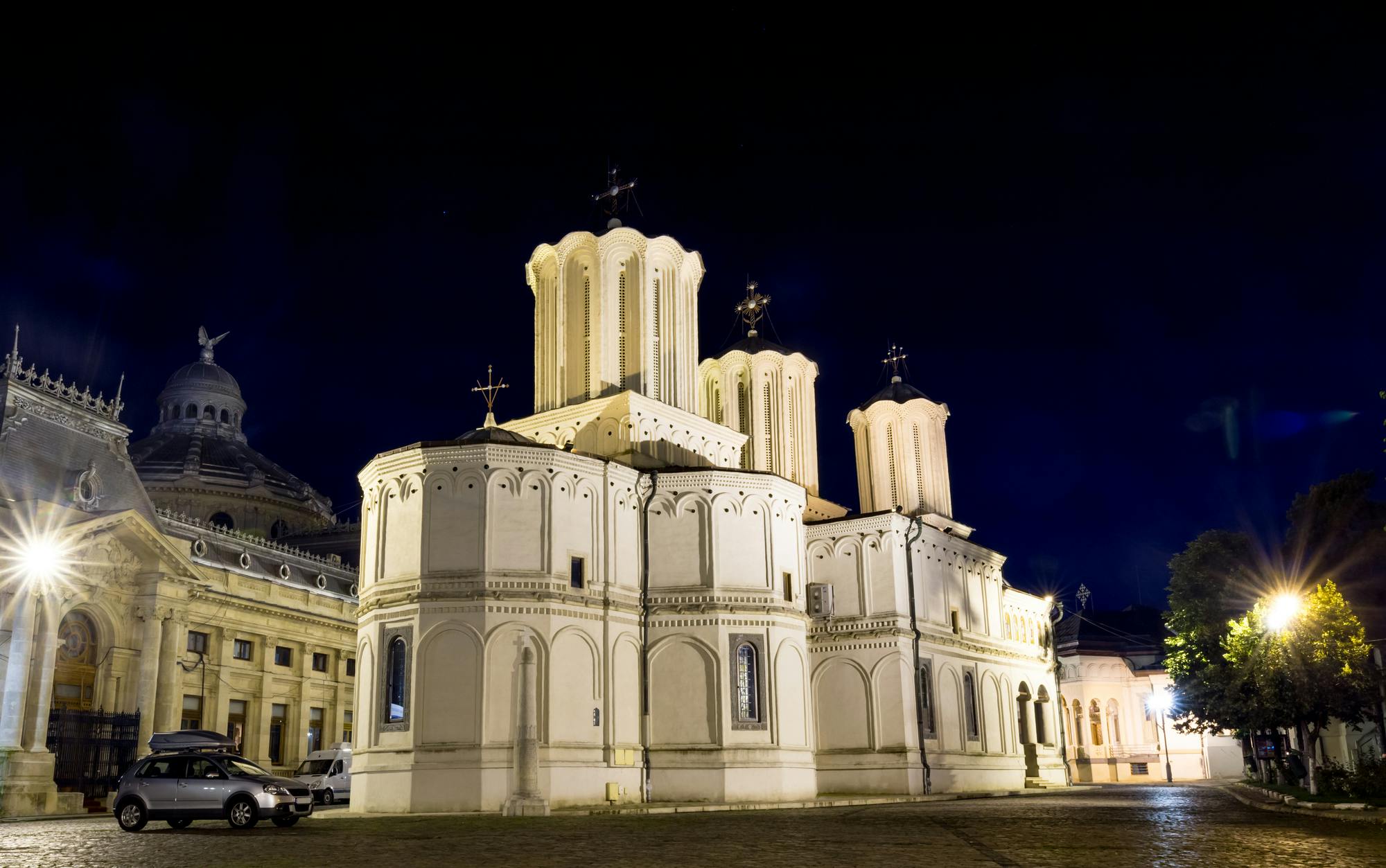 Illuminated white church with domes and crosses, set against a dark night sky. Nearby buildings and a parked van are visible.