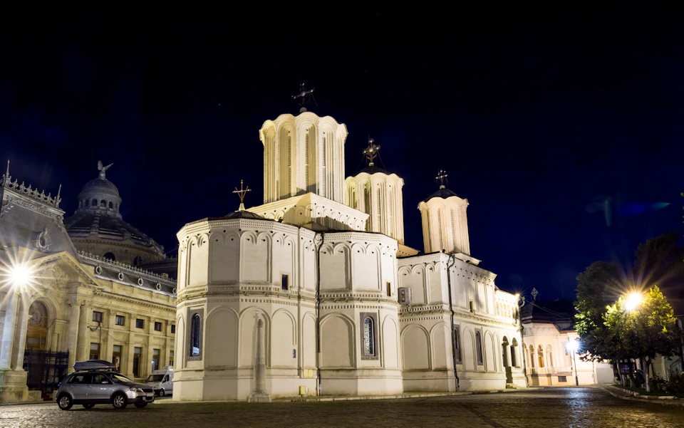 Illuminated white church with domes and crosses, set against a dark night sky. Nearby buildings and a parked van are visible.