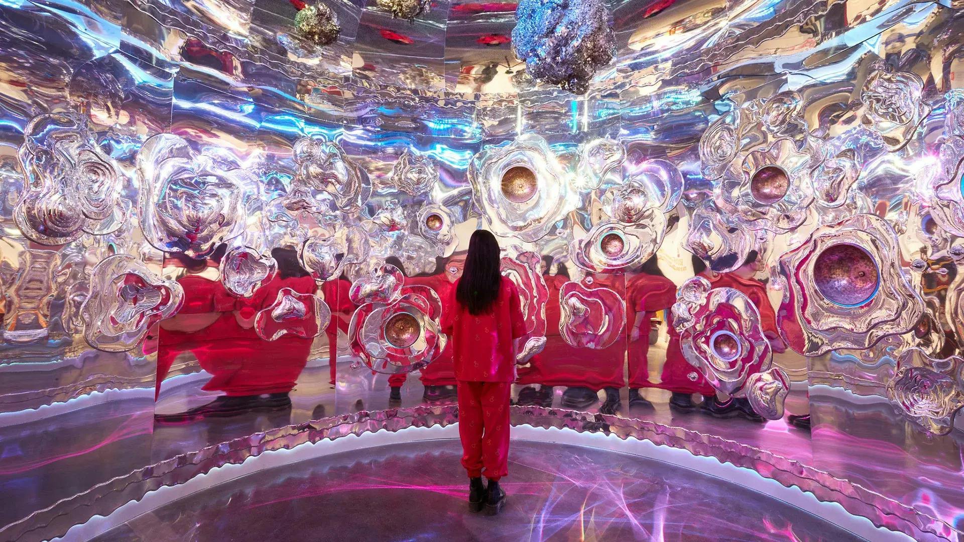 Osoba v červeném oblečení stojí uvnitř pohlcující, reflexní umělecké instalace s abstraktními, kovovými tvary na stěnách a stropě.