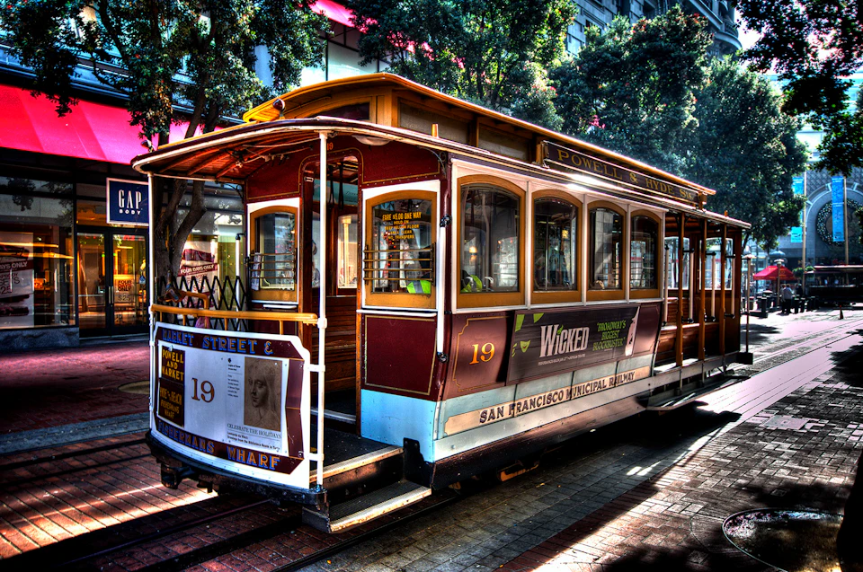 A red and white San Francisco cable car marked "Powell & Hyde" with advertising banners, parked on a sunlit street near trees and buildings.