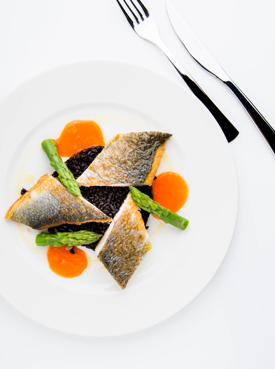 A plated dish featuring pan-seared fish fillets, arranged over black rice with asparagus spears, garnished with orange sauce.