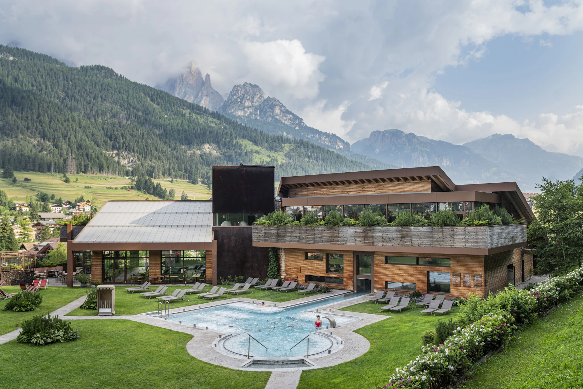 A modern building with a pool, surrounded by green grass and mountains under a cloudy sky.