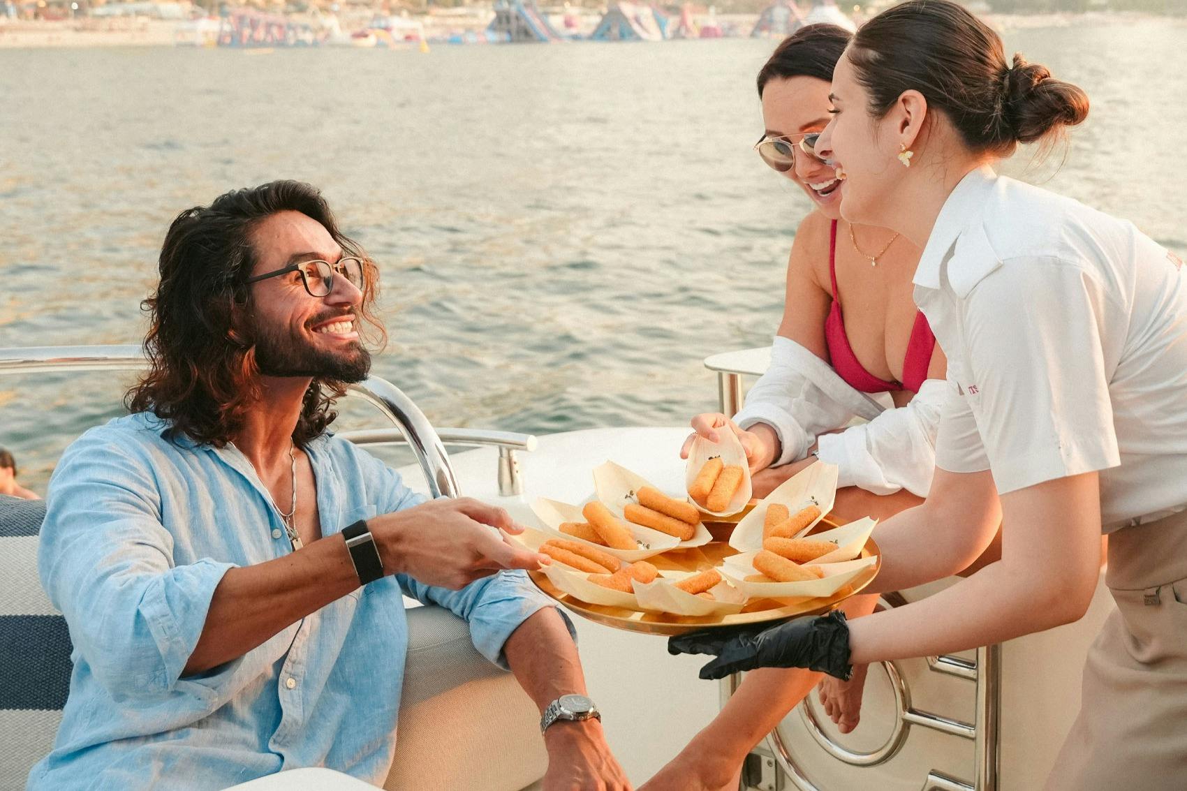 A man and two women on a boat enjoy snacks being served by one of them, with water and distant scenery in the background.