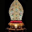 An ornate, jewel-encrusted golden mitre on a red, gold-trimmed base against a black background.