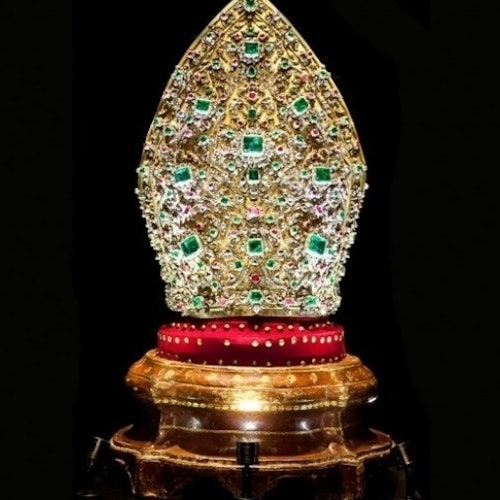 An ornate, jewel-encrusted golden mitre on a red, gold-trimmed base against a black background.