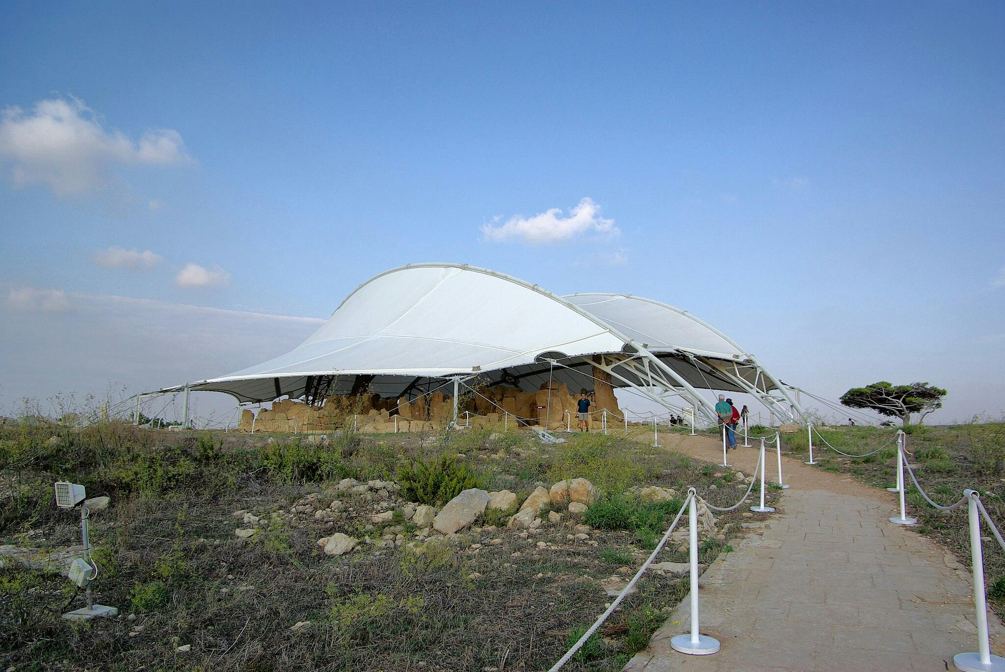 Bílá tahová konstrukce pokrývá archeologické naleziště s chodníkem lemovaným provazy, které k němu vedou, pod modrou oblohou s rozptýlenými mraky.