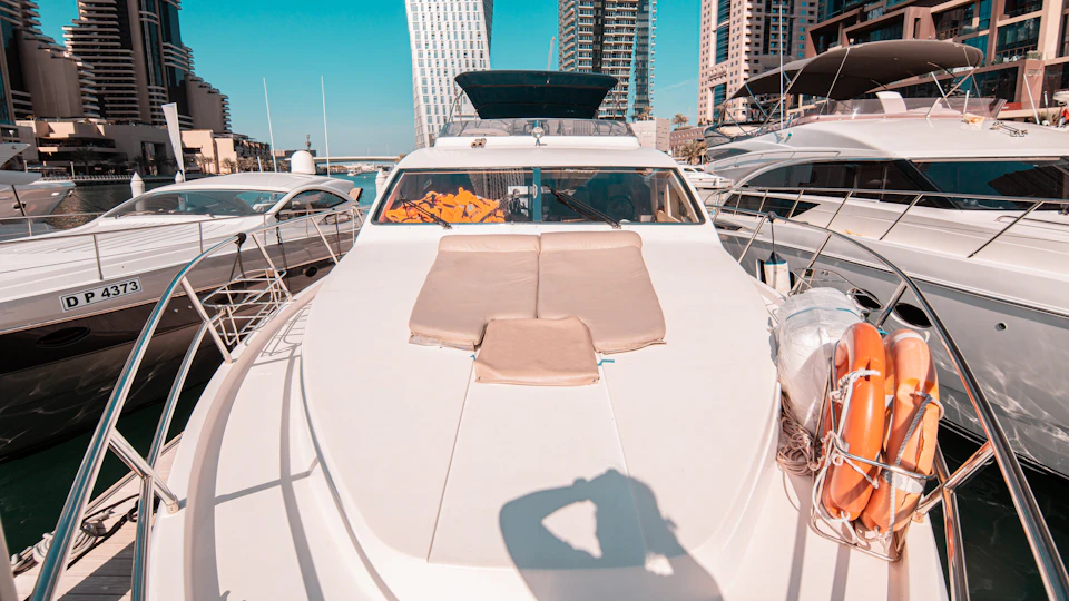 Luxury yacht docked in a marina, with beige cushions on the deck and city buildings in the background under a clear sky.