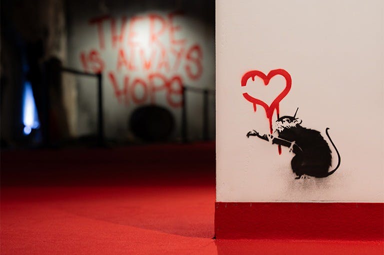 Stěna s graffiti uměním krysy držící štětec, malující červené srdce a text na pozadí, „VŽDY JE NADĚJE“.