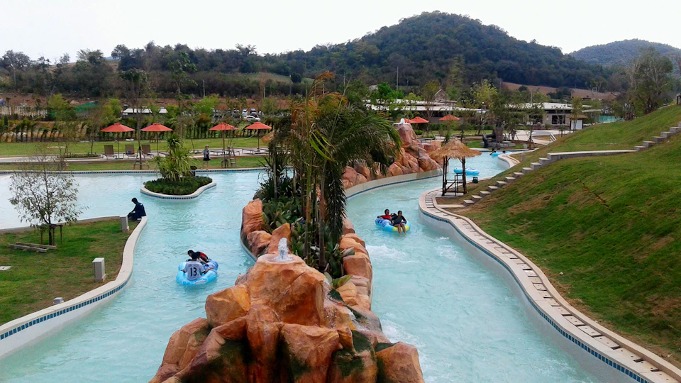 People float on tube rafts in a winding, artificial lazy river with rock formations and greenery. Hills and park structures in the background.