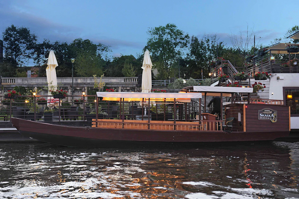 A boat with canopies moored by a riverside patio with umbrellas, stairs, and plants, set against a tree-lined background at dusk.