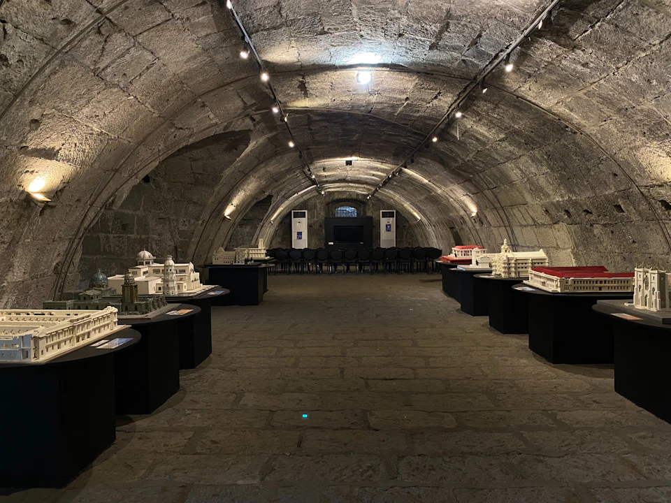 A vaulted stone room with architectural models on tables along the sides, and rows of chairs arranged at the far end.