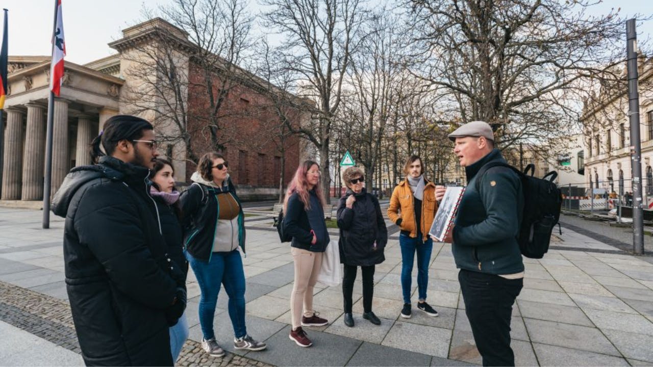A group of people stand near a historic building with flagpoles and leafless trees on a cloudy day.