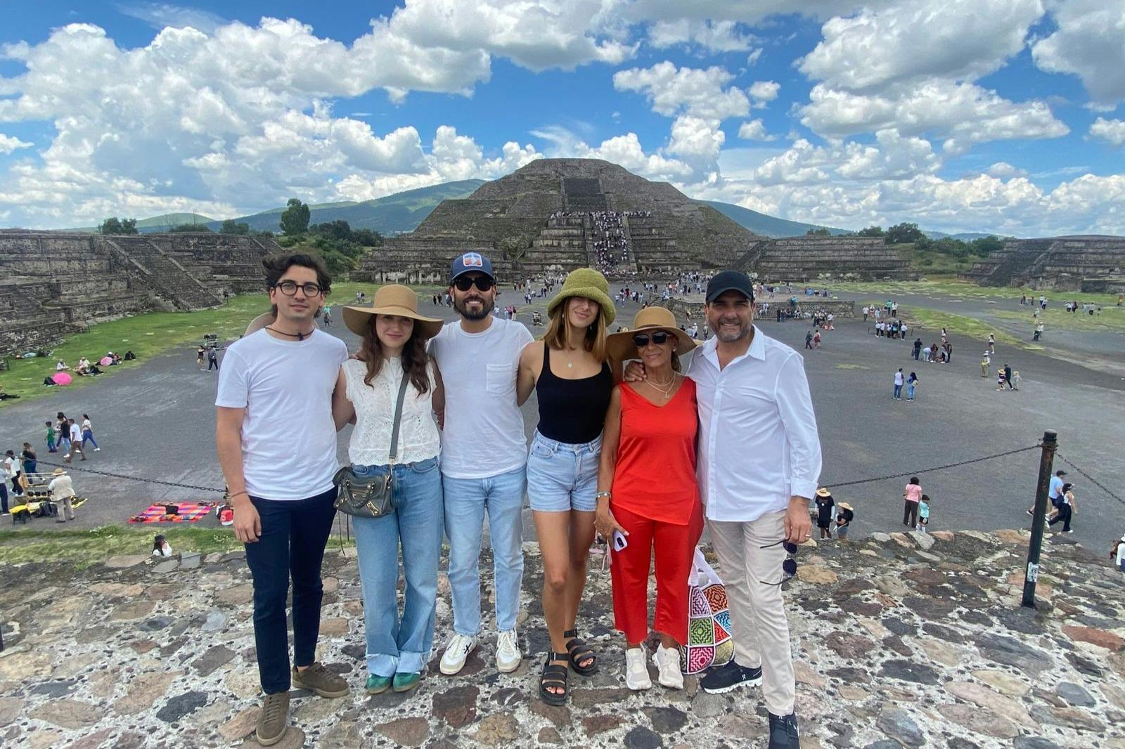 A group of six people standing on ruins with pyramids and other tourists in the background under a partly cloudy sky.