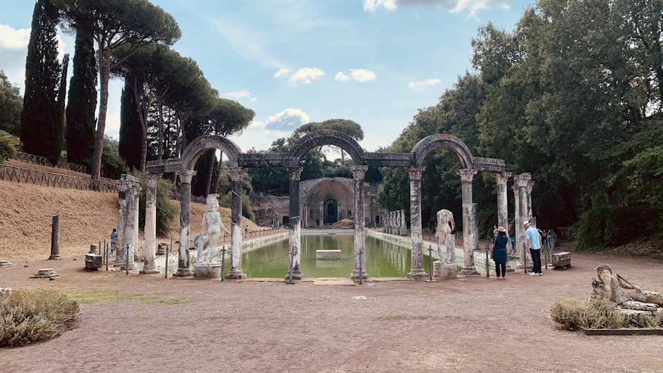 Ancient ruins with arched columns surrounding a rectangular pool. A person stands near the right edge, observing the scene.