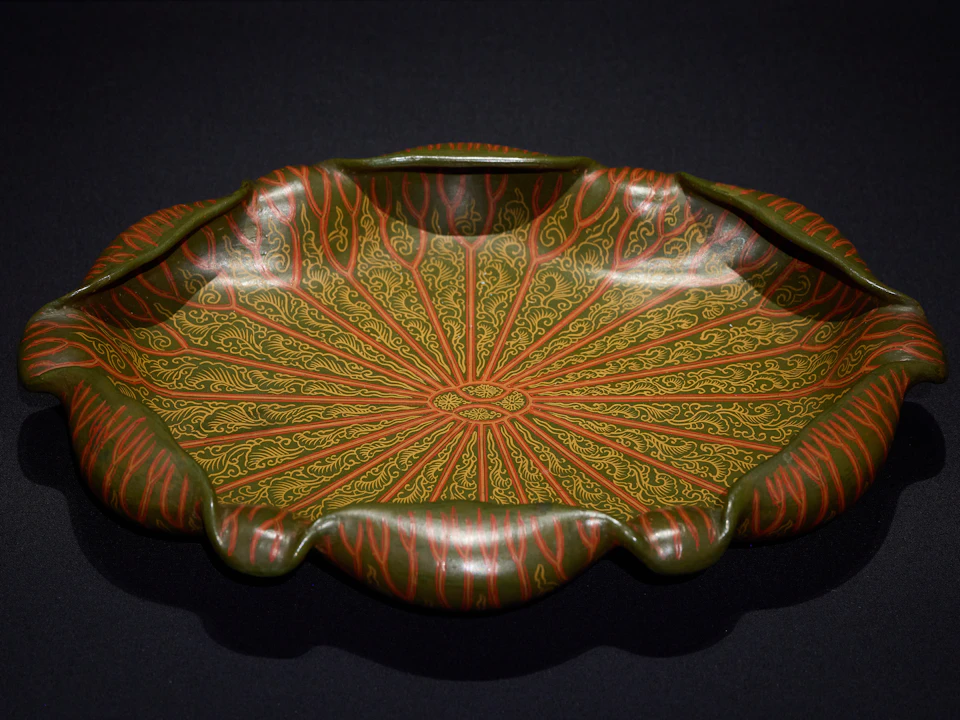 A decorative ceramic bowl with intricate red and gold designs on a green base, featuring a scalloped edge, set against a black background.