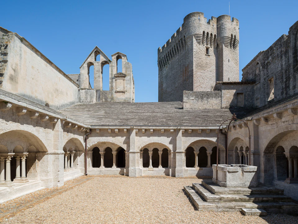 A medieval stone courtyard with arched walkways, a well in the foreground, and a castle tower in the background under a clear sky.