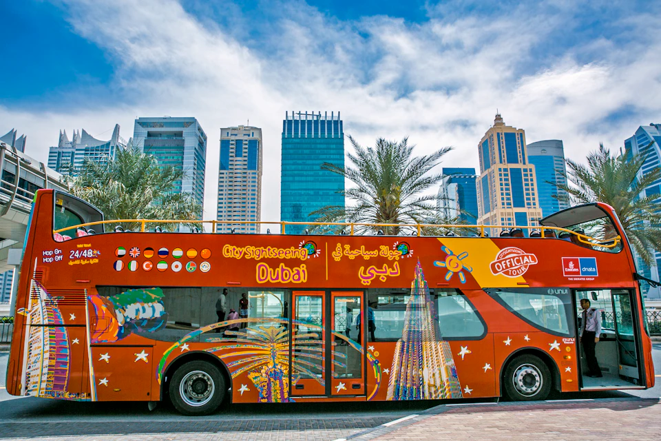 A colorful double-decker sightseeing bus in Dubai with skyscrapers and palm trees in the background under a partly cloudy sky.