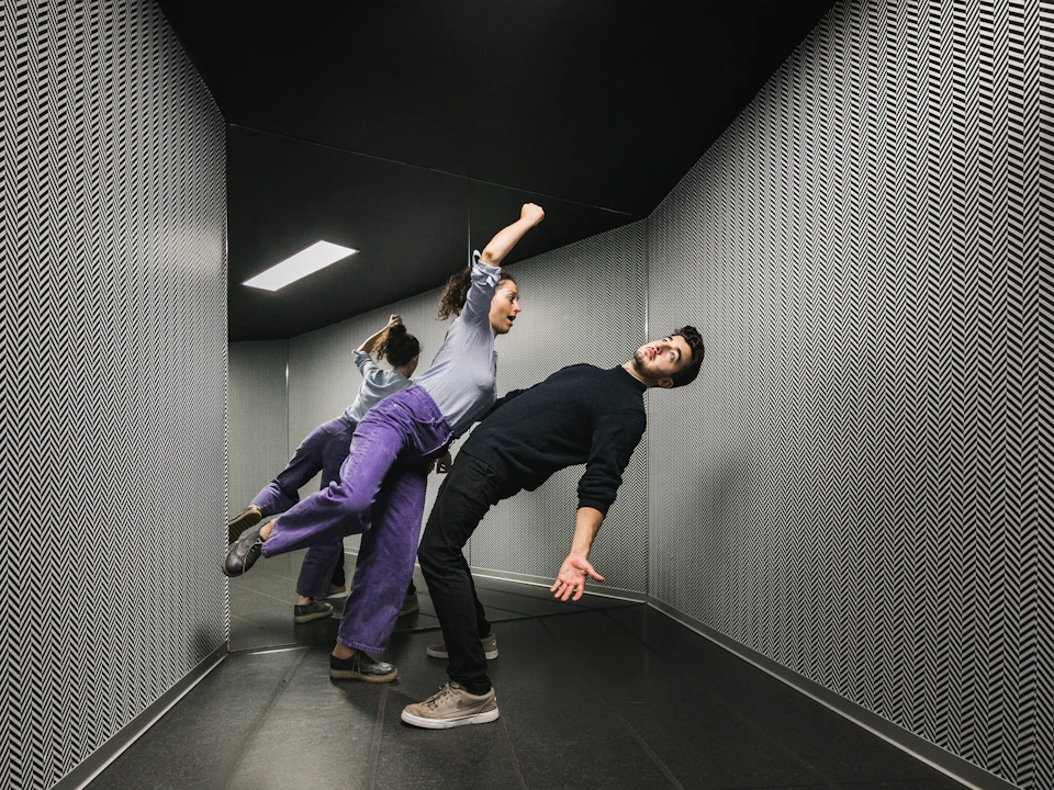 Two people appear to defy gravity, leaning at impossible angles in a mirrored, patterned room.