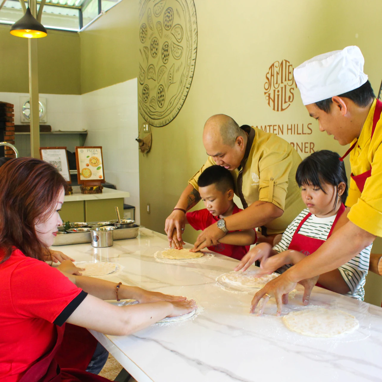 Children and adults in aprons making pizza dough on a marble counter under the guidance of instructors in a cooking class.