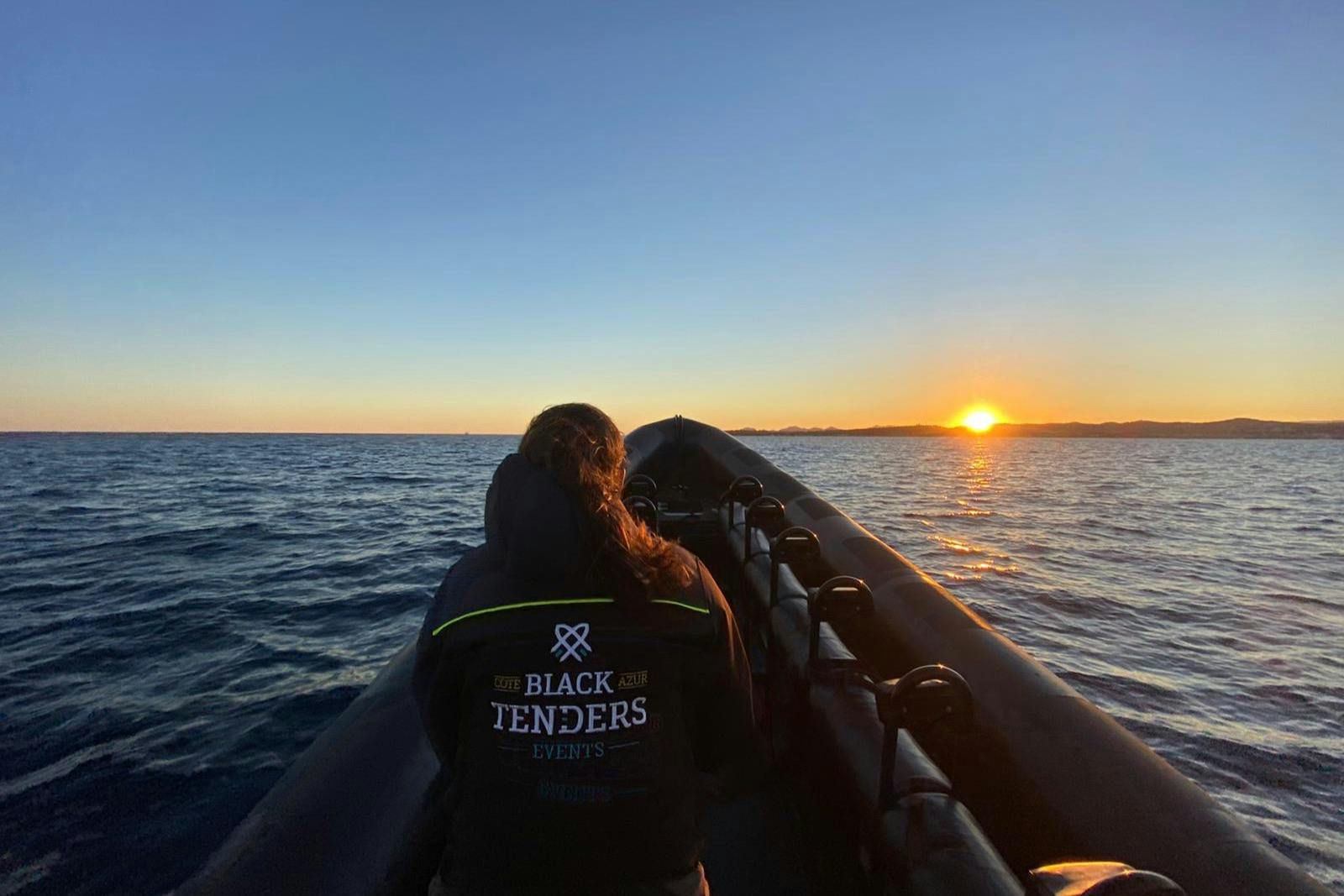 Person in a "Black Tenders Events" jacket sits at the front of a boat on the ocean at sunset.