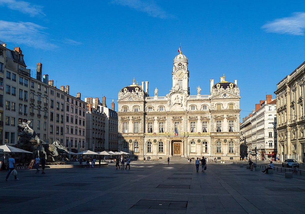 Chodci se procházejí po velkém náměstí s historickou ozdobnou budovou v pozadí pod jasně modrou oblohou.
