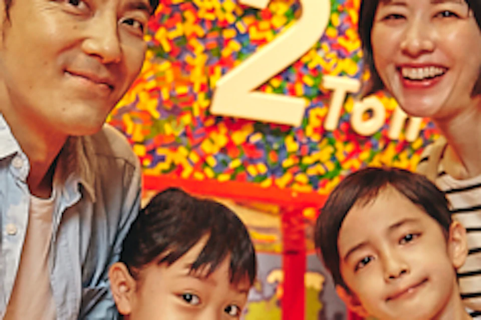 A family of four smiling inside a colorful, indoor play area with tubes, activities, and a large '2' visible in the background.