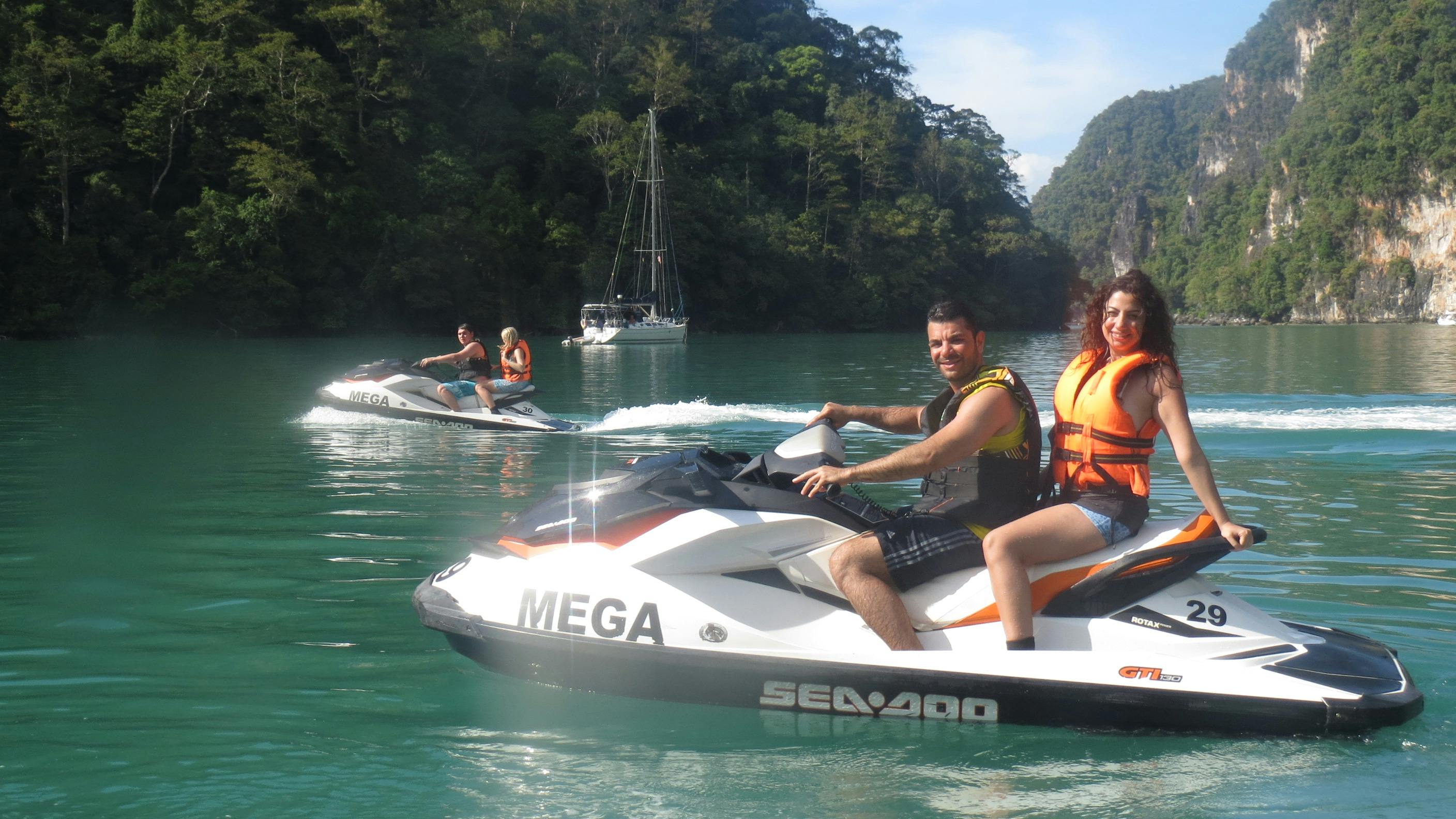 Un homme et une femme sur un jet ski dans une baie pittoresque avec des falaises et de la verdure en arrière-plan. Ils sont tous deux souriants et portent des gilets de sauvetage.
