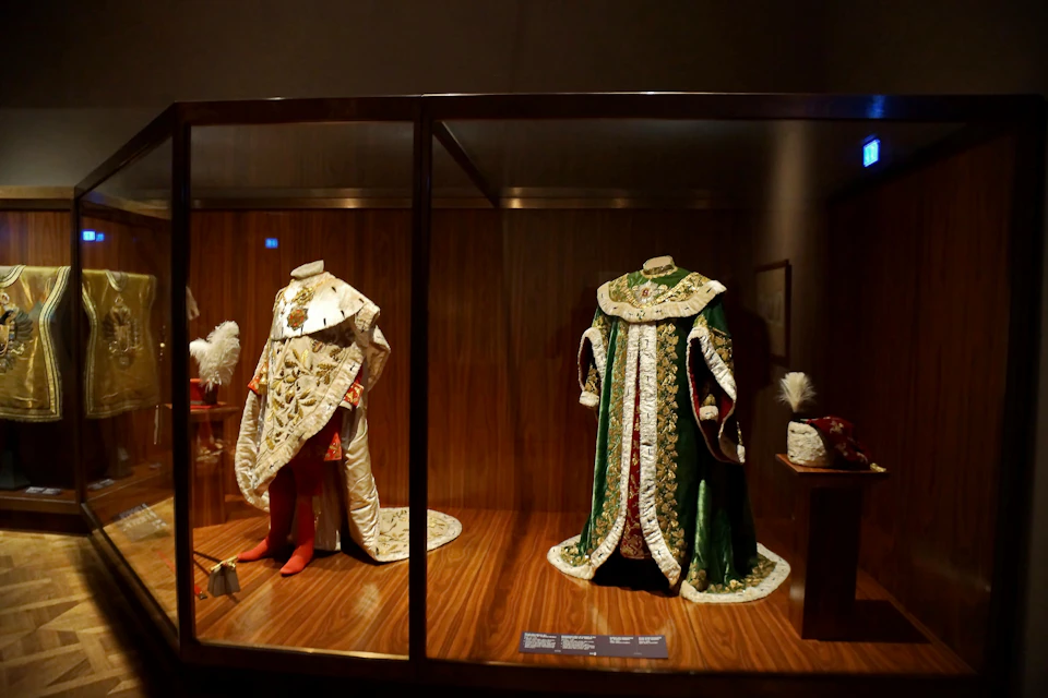 Two elaborate, headless royal garments displayed in a wooden glass case; one red and gold, the other green and gold.