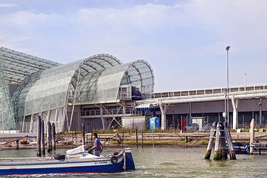 Person in a small motorboat on water near a modern building with large glass arches, against a background of an overcast sky.