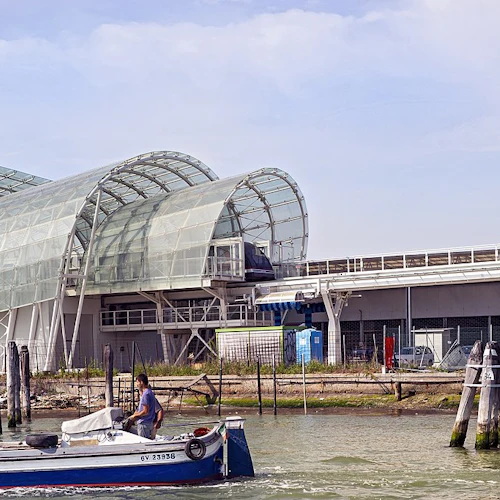 Person i en liten motorbåt på vatten nära en modern byggnad med stora glasbågar, mot en bakgrund av en mulen himmel.