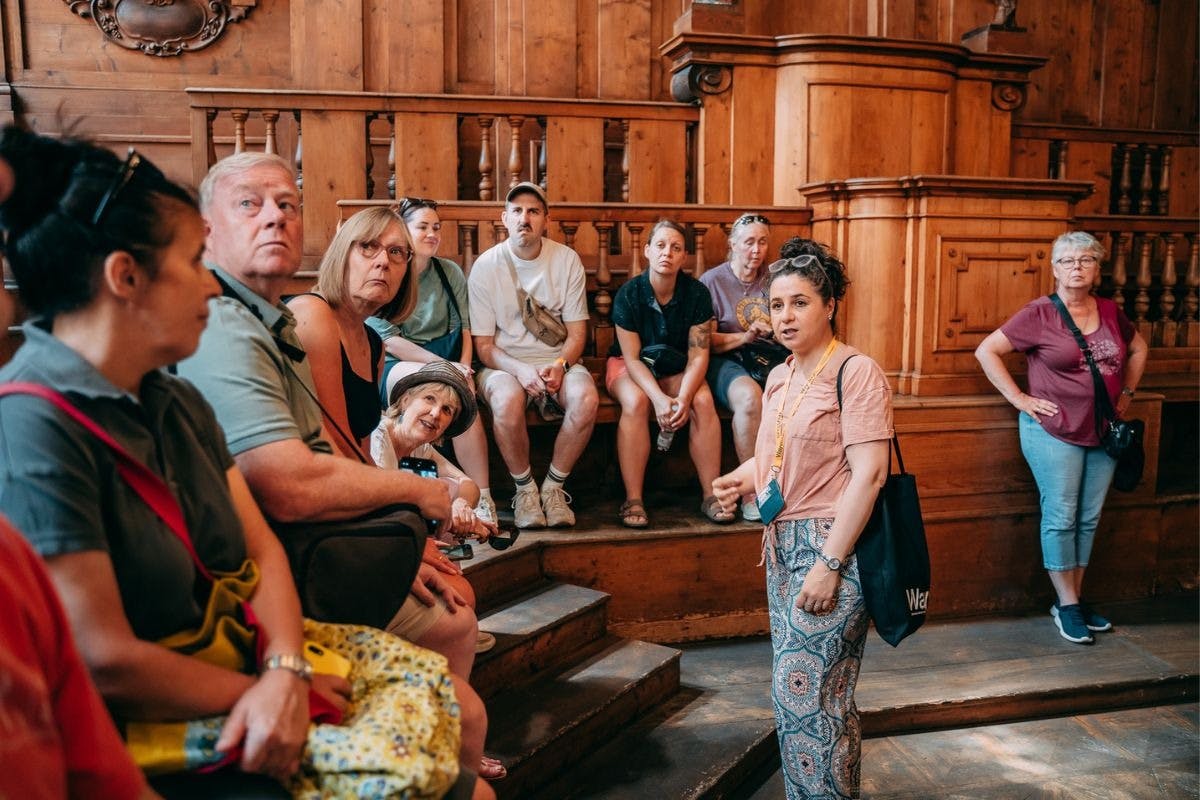A woman holding a guide tag speaks to a seated tour group inside a wooden historical building.
