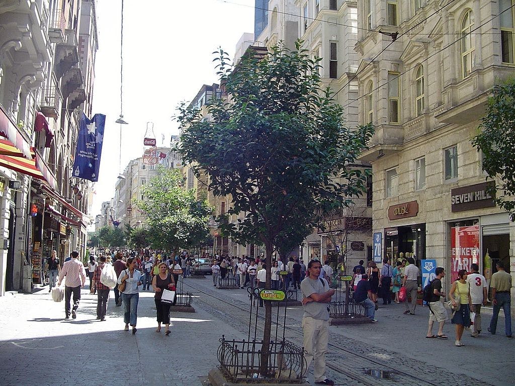 사람들이 걷고, 상점과 화려한 건물이 늘어서 있고, 중앙에 나무가 있는 번화한 거리.