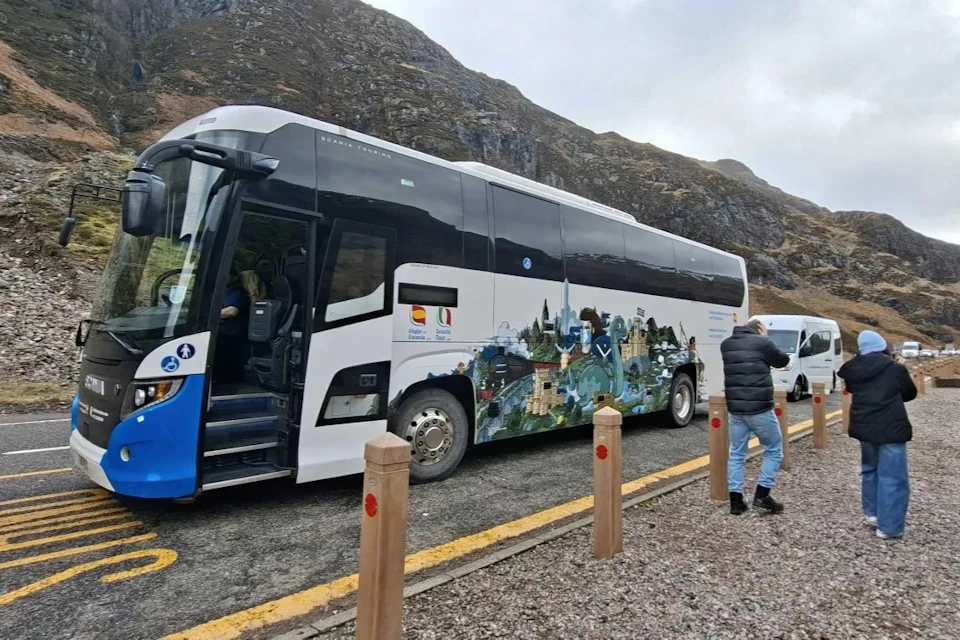 A bus with artistic imagery parked on a road near a mountainous landscape, with a person walking by.