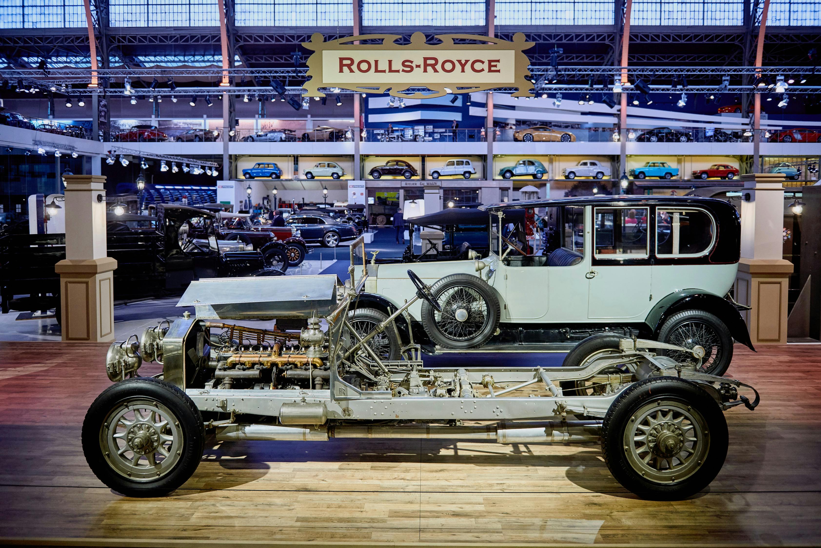 Deux voitures Rolls-Royce d'époque sont exposées, l'une montrant son châssis et son moteur, avec un panneau "Rolls-Royce" bien visible au-dessus.