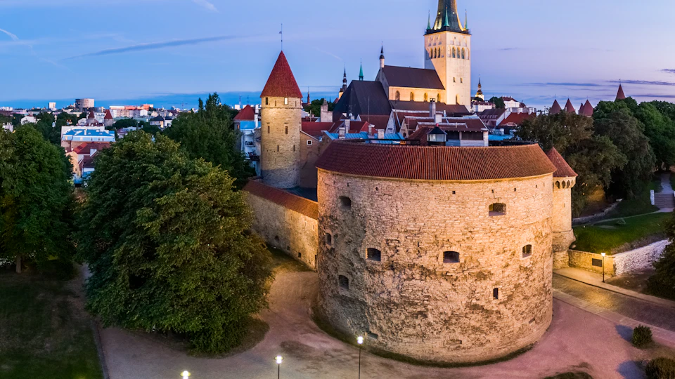 Medieval stone towers and walls beside lush trees, with a church spire and rooftops in the background during twilight.