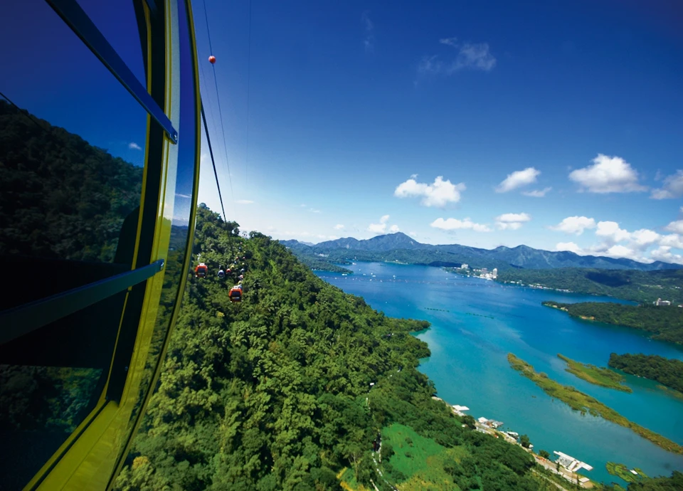 A cable car ascending over a lush green landscape with a large blue lake and distant mountains under a clear sky.