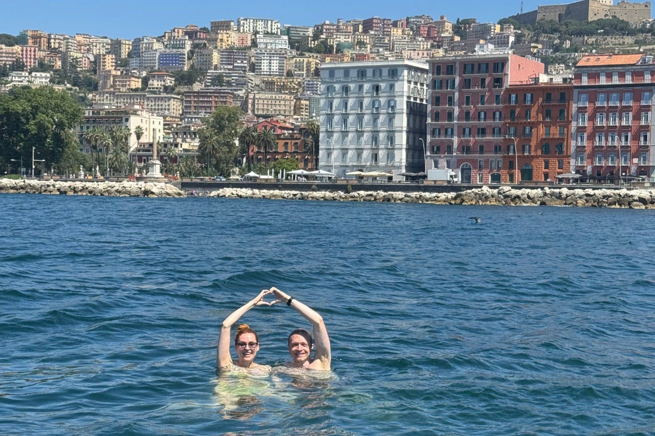 Dos personas sonriendo y formando un corazón con las manos mientras nadan en el mar, con un paisaje urbano de fondo.