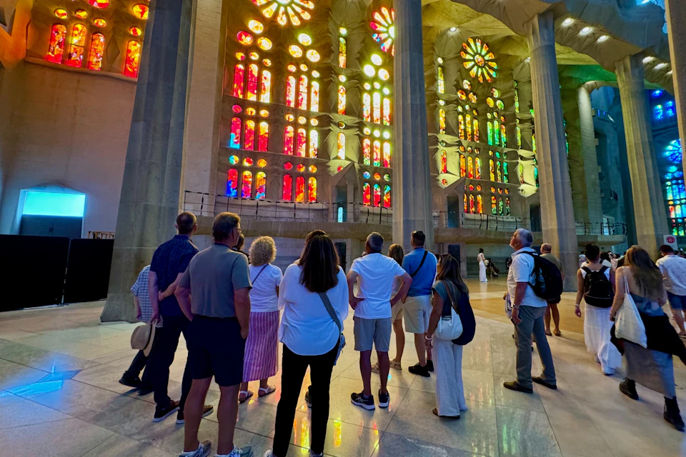 A group of people stands inside a cathedral, admiring colorful stained glass windows illuminated by sunlight.
