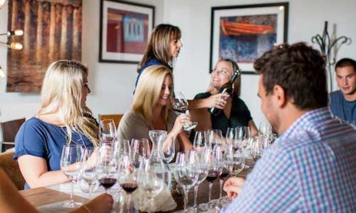 Lidé sedí kolem stolu s sklenicemi na víno, některé plní žena nalévající víno, v neformálním, vnitřním prostředí.