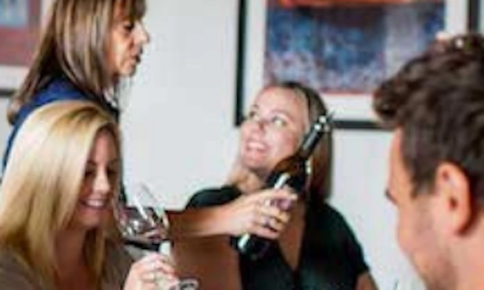 People sitting around a table with wine glasses, some being filled by a woman pouring wine, in a casual, indoor setting.