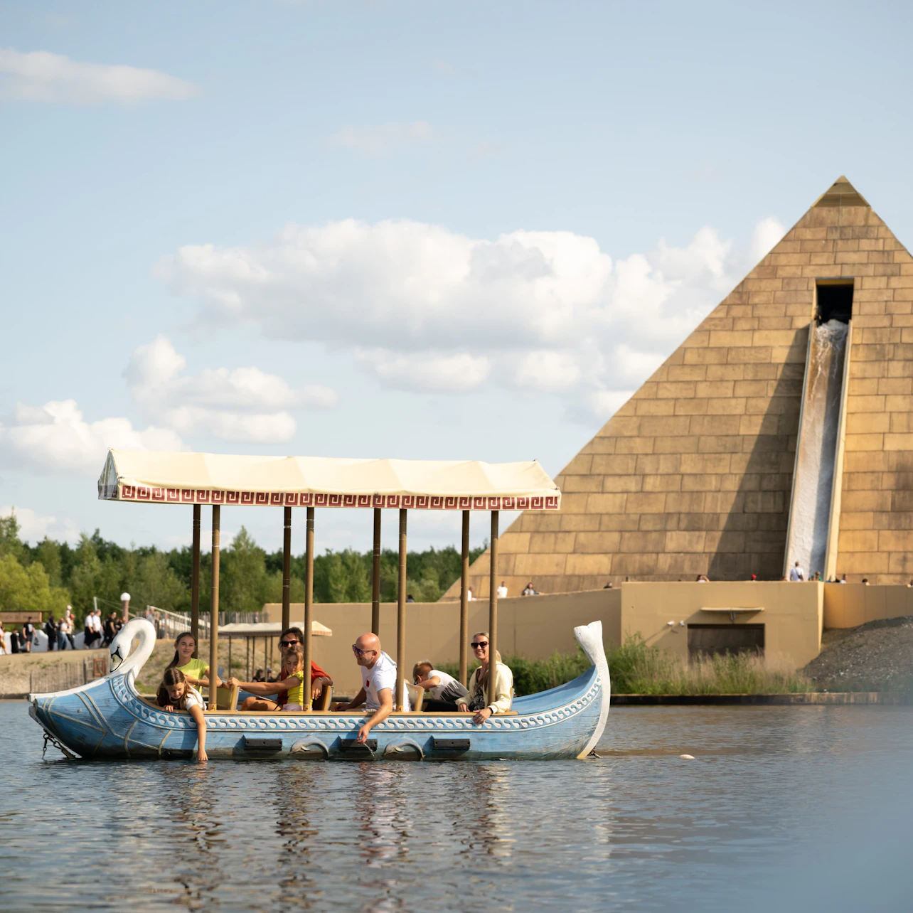 A group of people ride a decorated boat on a waterway near a large pyramid structure with a waterfall feature.